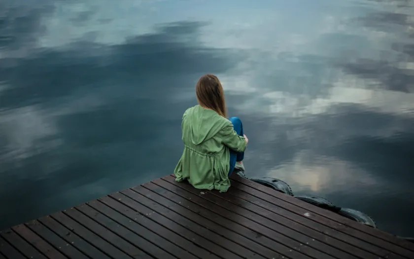 Imagem de uma mulher de costas, pensativa, junto a um lago. Crédito: Keenan Constance/Canva