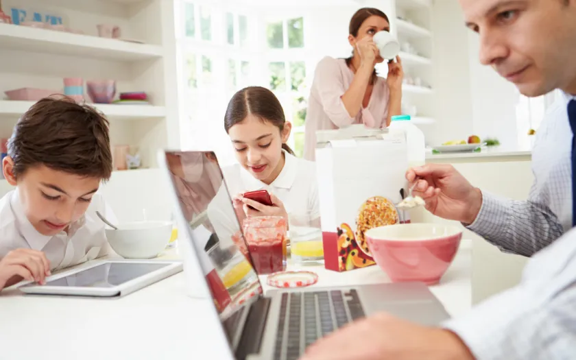 Uma família toma o pequeno-almoço na cozinha, mas o pai e os dois filhos estão a ver ecrãs de computador ou telemóvel. Crédito: Canva