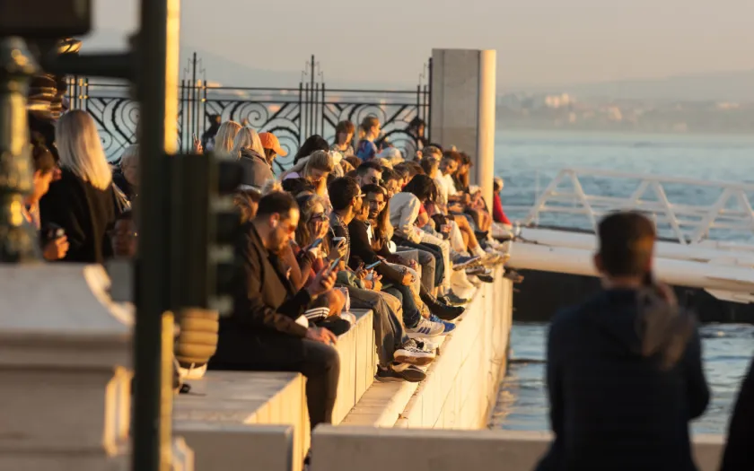 Pessoas de várias idades estão sentadas junto ao rio tejo, em Lisboa, ao entardecer. Crédito: Shutterstock
