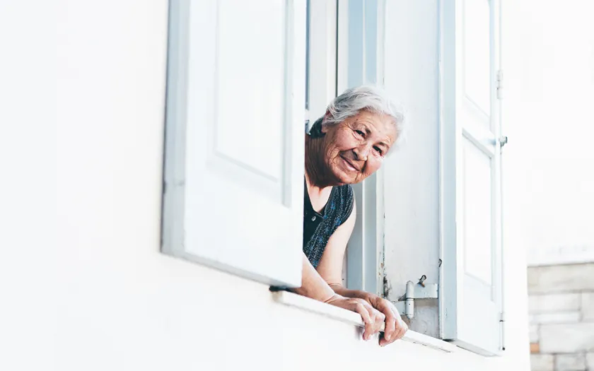 Uma mulher idosa e sorridente está à janela a espreitar para a rua, num dia de verão. Crédito: Mike Jones (Canva) 