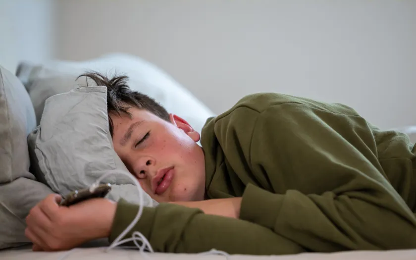 um adolescente dorme com o telemóvel na mão, sob um fundo neutro. Crédito: Canva.