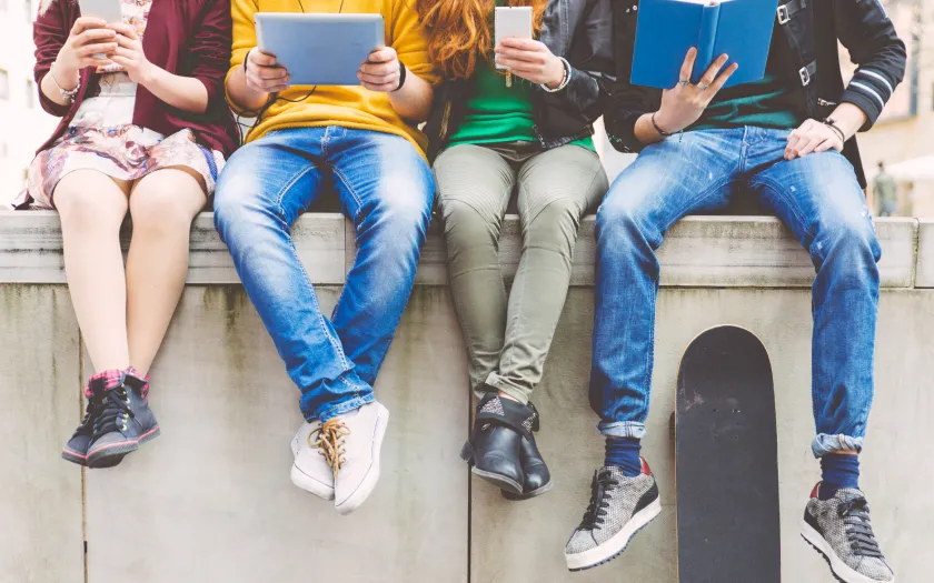 vários adolescentes estão a ver conteúdos no telemóvel e ipad, sentados num muro, num ambiente citadino. Crédito: Canva