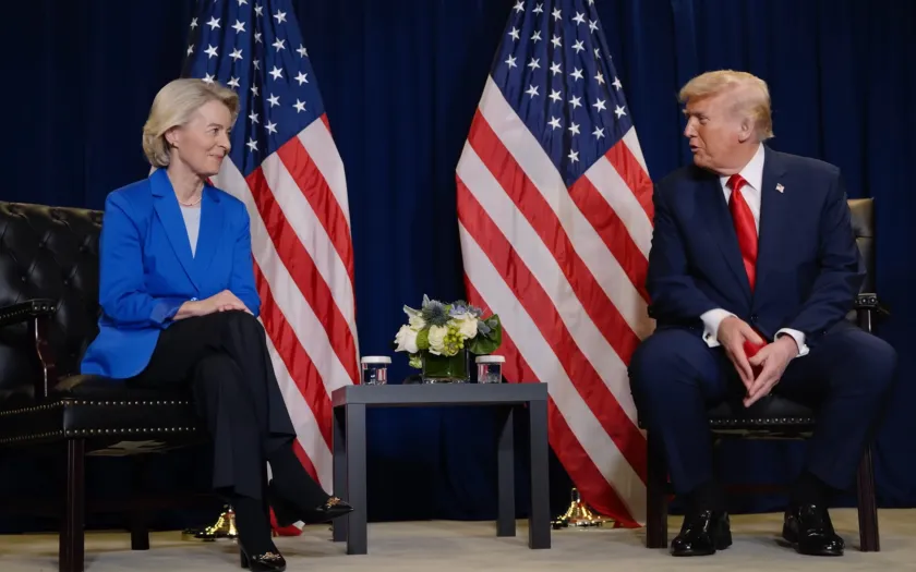 Ursula von der Leyen sentada ao lado de Donald Trump, com duas bandeiras dos EUA atrás.