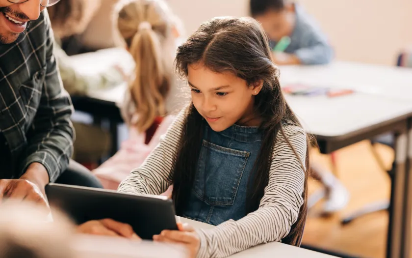 Uma menina de cabelos escuros, com um tablet na mão, tendo como fundo uma sala de aula. Crédito: Shutterstock