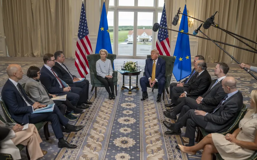 Ursula von der Leyen sentada ao lado de Donald Trump, com bandeiras da UE e dos EUA.