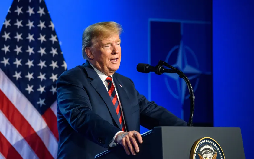 Donald Trump a falar, num púlpito. Fundo azul, com uma bandeira dos EUA e o símbolo da Nato. Créditos: Gints Ivuskans/shutterstock