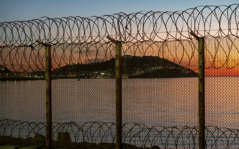Ceuta, cidade espanhola no norte da África, vista do Marrocos atrás de uma cerca de arame espinhoso localizada na fronteira marroquina, contra um deslumbrante pôr do sol sobre o mar. Crédito: Shutterstock