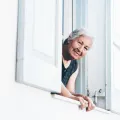 Uma mulher idosa e sorridente está à janela a espreitar para a rua, num dia de verão. Crédito: Mike Jones (Canva) 