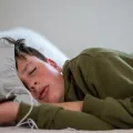 um adolescente dorme com o telemóvel na mão, sob um fundo neutro. Crédito: Canva.