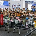 Imagem de robôs, dentro de um ringue, com luvas de boxe. Uma plateia a assistir e a filmar.