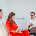 Três pessoas sentadas num palco. Dois homens de fato, uma senhora com um vestido vermelho. Ao fundo, o logotipo da Fundação.
