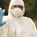 Imagem de uma pessoa, vestida com um fato de segurança branco, máscara, óculos e luvas azuis.