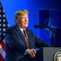 Donald Trump a falar, num púlpito. Fundo azul, com uma bandeira dos EUA e o símbolo da Nato. Créditos: Gints Ivuskans/shutterstock