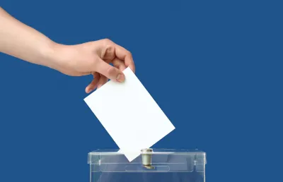 Imagem de uma mão a depositar o voto numa urna transparente, sob um fundo  azu. Crédito: Shutterstock.