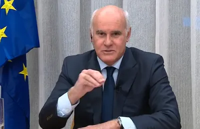 Imagem de um homem, de fato e gravata a falar, sentado numa secretária e tendo uma bandeira da União Europeia ao lado. D/R.