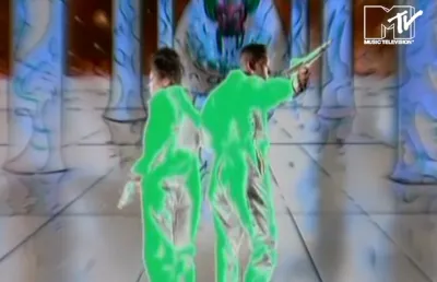 Imagem de um casal de costas, vestido com um fato verde fluorescente, num ambiente espacial.  D/R.