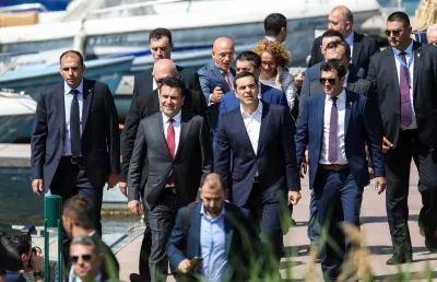 Vários de homens de meia idade, vestindo fatos , caminham pelas ruas.  A comitiva é  encabeçada pelo primeiro-ministro grego, Alexis Tsipras, e pelo seu homólogo macedónio, Zoran Zaev, que se preparam para a assinatura do acordo relativo ao novo nome da Macedónia, na aldeia de Psarades. Crédito: Shutterstock.