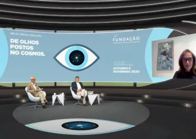 Carlos Fiolhais e outro homem sentados, num estúdio de televisão, a olhar para uma tela, na qual Ann Druyan está a falar.