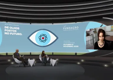 Um homem e uma mulher sentados, num estúdio de televisão, a olhar para uma tela, na qual está Elissa Epel a falar. Do lado oposto da tela lê-se "De olhos postos no futuro". 