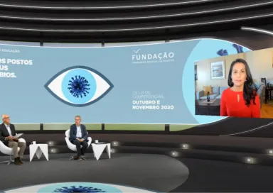 Dois apresentadores sentados, num estúdio, a olhar para uma tela na qual se vê Nahid Bhadelia a falar