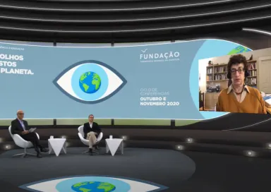 Entrevista virtual, com dois homens sentados em cadeiras num estúdio, e a historiadora de Ciência Naomi Oreskes a falar, numa tela, com o texto "DE OLHOS POSTOS NO PLANETA" e o logotipo da Fundação.