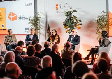 Uma plateia sentada, de costas, a olhar para um palco onde estão quatro oradores, entre eles Pedro Passos Coelho e Elvira Fortunato.