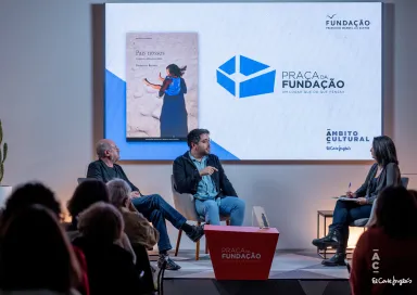 Uma plateia, de costas, a olhar para um palco, onde estão dois homens sentados a falar.