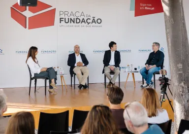 Quatro pessoas em palco, sentadas em cadeiras pretas, a falar com plateia a assistir. Logo e texto "Praça da Fundação".