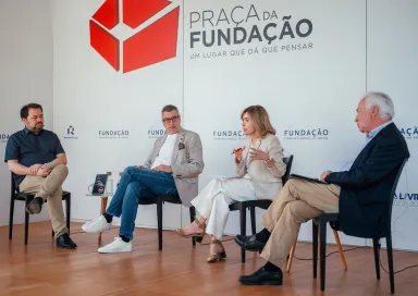 Quatro pessoas sentadas em cadeiras pretas, a debater entre si. Ao fundo, o texto "Praça da Fundação"