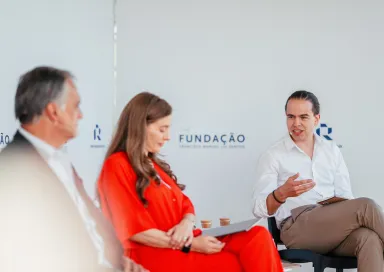 Três pessoas sentadas num palco. Dois homens de fato, uma senhora com um vestido vermelho. Ao fundo, o logotipo da Fundação.