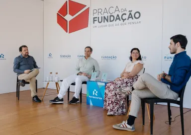 Quatro pessoas sentadas num palco, em cadeiras pretas, a apresentar um livro que está pousado em cima de uma mesa. Fundo com logotipos da Fundação.
