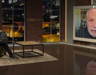 Um homem sentado num estúdio de televisão e outro num ecrã, durante uma entrevista.
