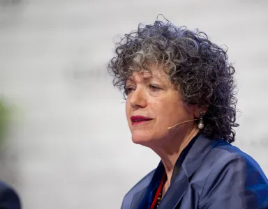 A professora Vivien A. Schmidt sentada, com um microfone sem fios, sobre um fundo branco. 