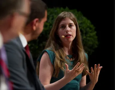 Mulher a falar num painel, a gesticular, e com um microfone na orelha. Fundo com plantas.