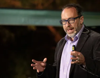 O fundador da Wikipedia Jimmy Wales a gesticular enquanto discursa em palco, sobre um fundo escuro.