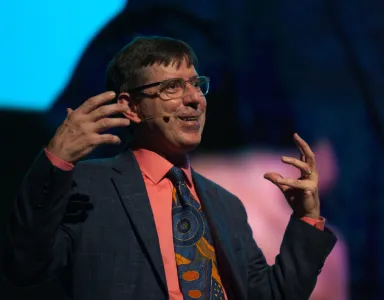 Homem de óculos em palco, gesticulando com entusiasmo. Veste uma camisa cor de salmão.