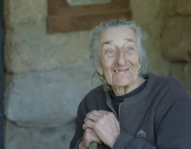 Retrato de uma mulher idosa, com as mãos apoiadas no cimo do cabo de uma vassoura. Atrás, uma parede de pedra.