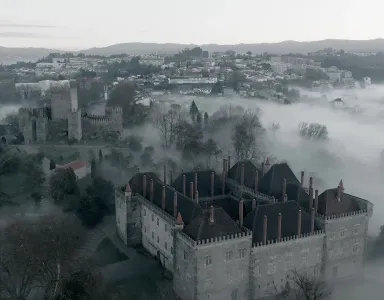 Castelo envolto em névoa, com vista para uma cidade nas colinas ao fundo.