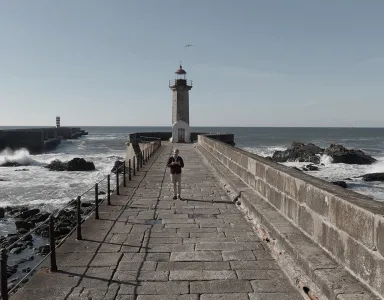 Homem a caminhar num caminho de pedra, rumo a um farol, junto ao mar agitado.