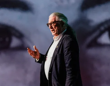 Howard Williamson, professor de Política Europeia, a falar de pé, gesticulando, sobre um fundo de dois olhos gigantes.