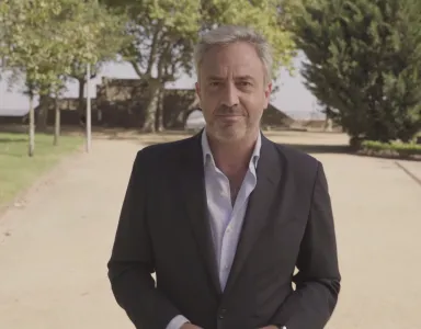 Homem de fato sorridente, cercado por árvores e vegetação. Céu limpo ao fundo.