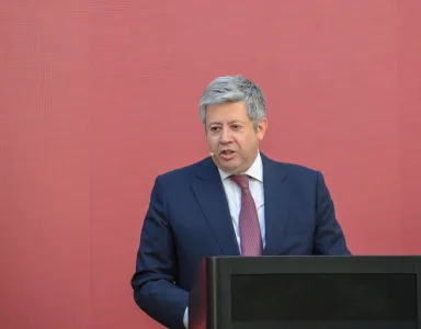 José Soares dos Santos, vestido de fato escuro, camisa branca e gravata vermelha, a discursar. Fundo rosa.