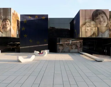Edifício moderno com rostos de várias pessoas e estrelas da União Europeia nas fachadas.