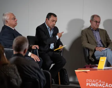Três homens sentados em cadeiras, um deles gesticula. Texto: "PRAÇA DA FUNDAÇÃO" em destaque numa mesa laranja.