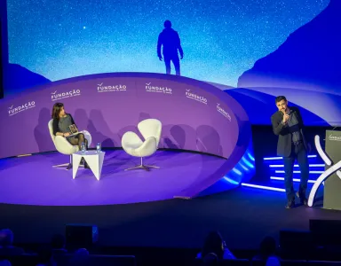 Imagem de um palco bem iluminado, com duas cadeiras brancas, um apresentador e texto "FUNDAÇÃO" ao fundo.