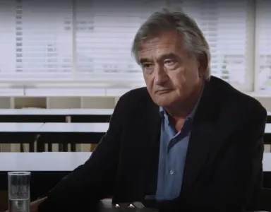 Homem de cabelo grisalho, Antony Beevor, sentado numa mesa vazia, copo de água ao lado. Ambiente escolar.