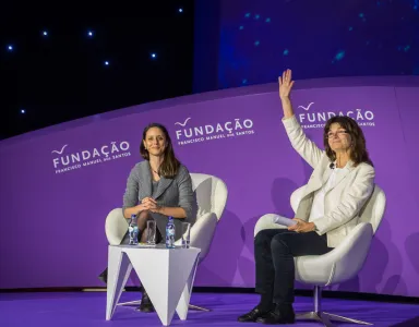 Duas mulheres sentadas, num palco com fundo roxo, uma delas está a acenar. Logo "FUNDAÇÃO".