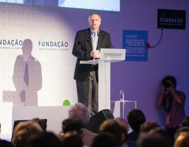 O professor Dani Rodrick a discursar, num púlpito, para uma plateia. Fundo com o logo da Fundação.