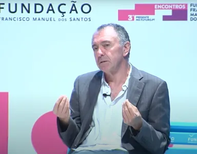 Um homem sentado, de fato cinzento e camisa branca, fala e gesticula. Logotipo "Fundação" sobre um fundo claro.