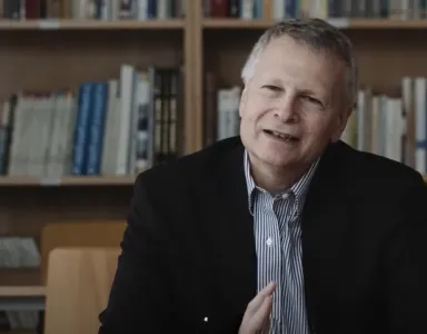 O economista político Dani Rodrik, de cabelo grisalho, sorrindo, sentado à mesa com prateleira de livros ao fundo.